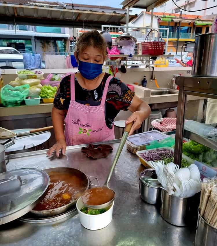 A lady making laksa - ladling fresh broth into a bowl 