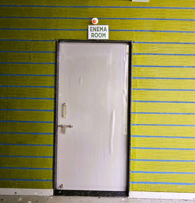 Shows the door of the Enema Room with Enema Room written above and a red light 