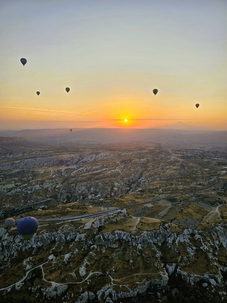 taken from high up looking down at the ground with lots of balloons in the distance and sun rising in Cappadocia