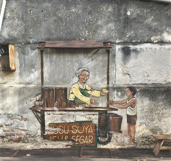 A mural showing an old fashioned stall with a lady selling soya milk to a young boy.