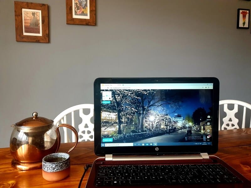 A laptop on a wooden table, on the screen there are cherry blossoms by a road. Next to the laptop is a small Japanese style cup and pot of tea. Behind the laptop is a grey wall, with some pictures, one is of Madam Butterfly.