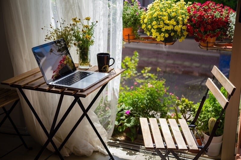 An image of a laptop on a table next to an open window, with lots of flowers and a linen curtain swaying in the breeze