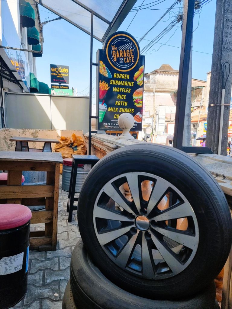 Outside of The Garage, shows a large wheel and industrial looking chairs and tables. A ign reads burger, submarine, rice, kottu, milk  shakes