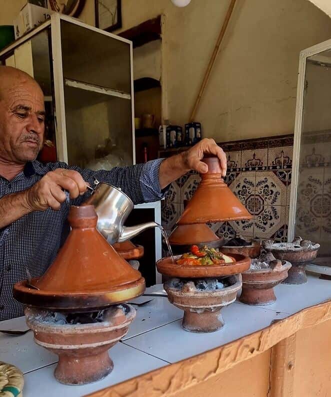 Man pouring water into tagines cooking on charcoal burners in in Taroudant city. 